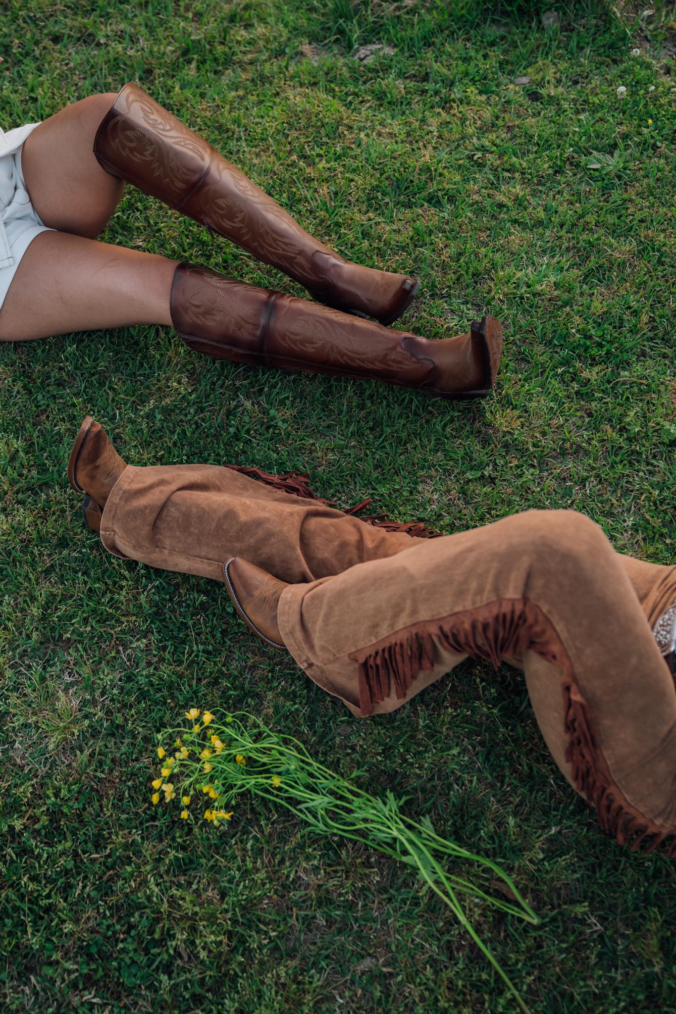 Two women's pair of legs laying on grass, showcasing Rock'em boots.