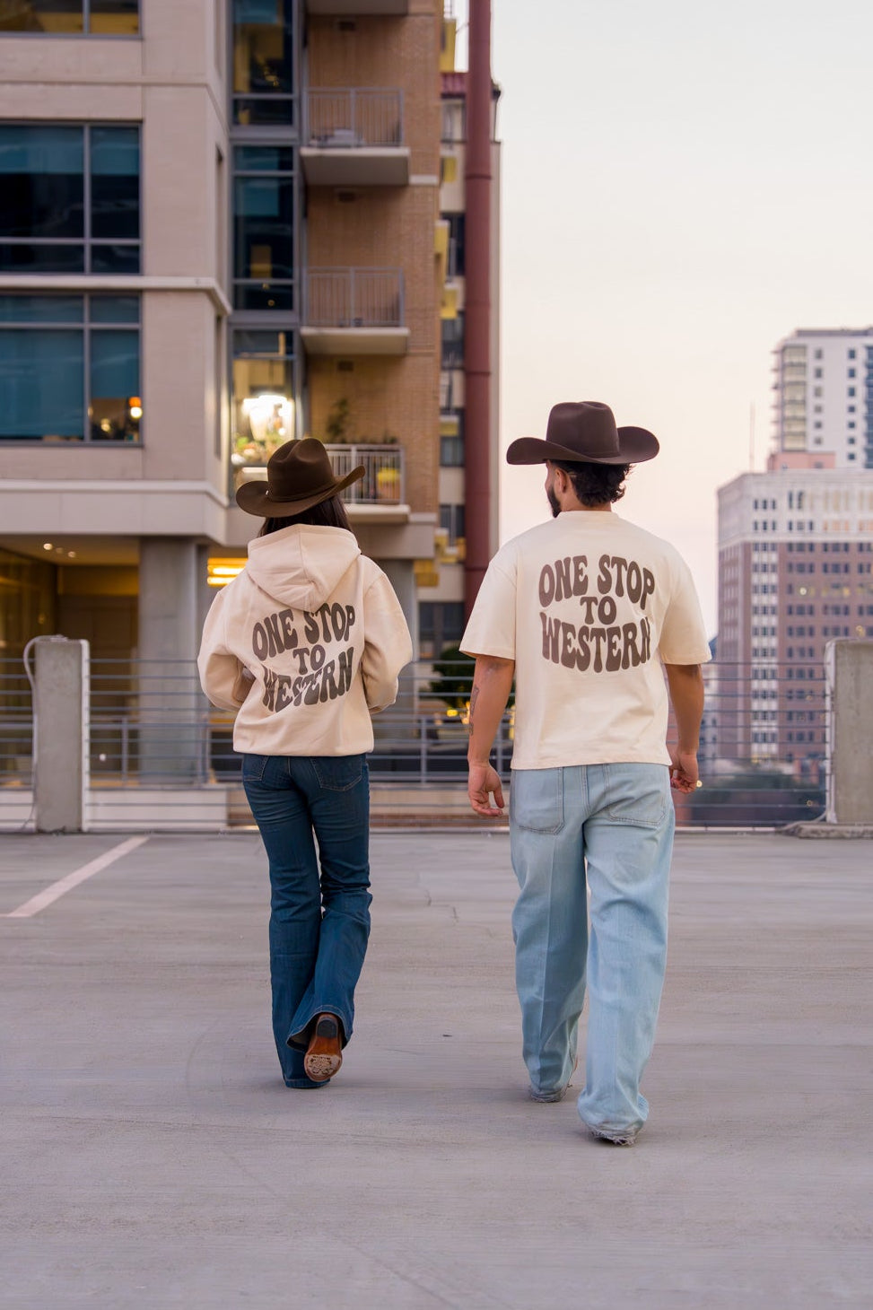 Two people wearing cowboy hats and 'One Stop to Western' shirts walking on a rooftop with cityscape in the background.