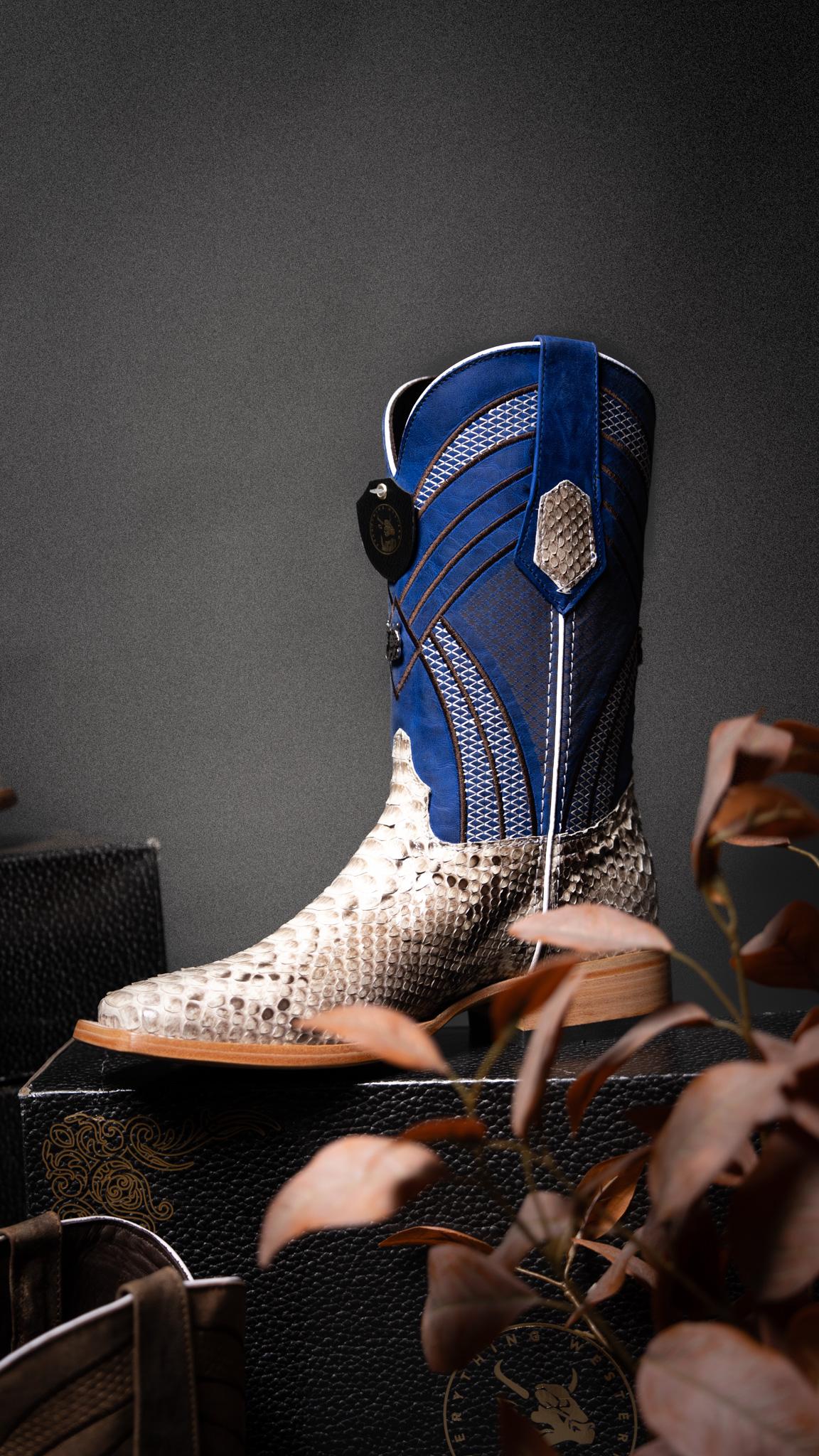 A western-style cowboy boot with a blue shaft and a beige-and-white snakeskin-patterned foot, displayed on a dark surface with decorative leaves in the foreground.