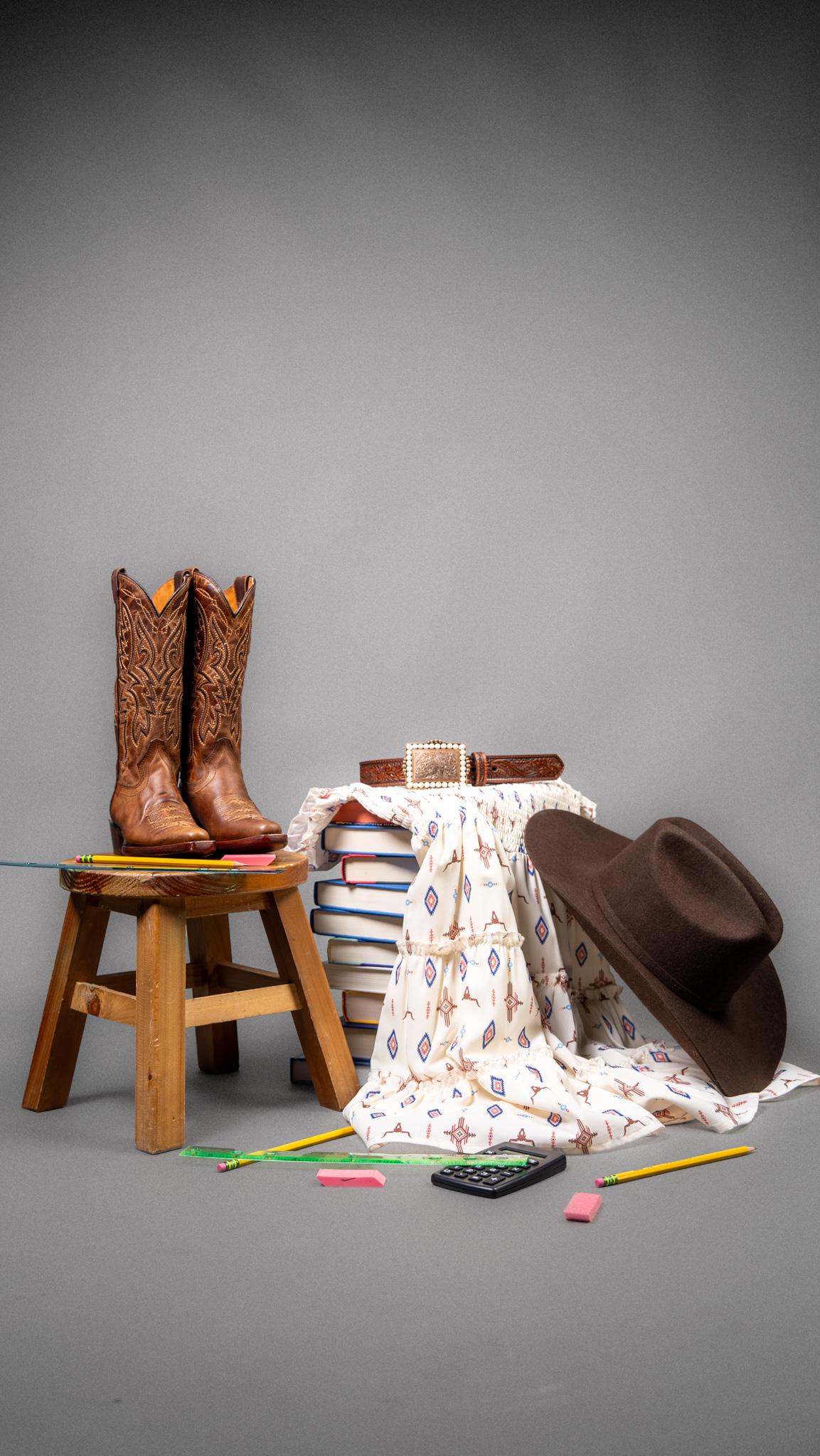 A styled studio still life featuring a pair of brown cowboy boots on a wooden stool, a dark brown wide-brim hat leaning against a stack of patterned books and a white patterned cloth, with scattered colored pencils and a calculator on a gray background.