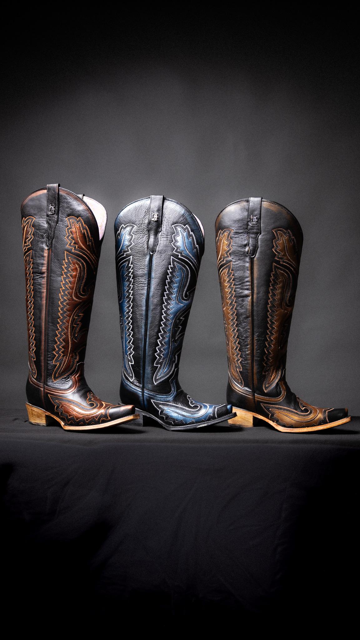 Three tall western cowboy boots displayed side by side on a dark surface against a dark gray background. The boots are shown in three colorways: brown with tan and black western embroidery, black with blue western embroidery, and brown with black western embroidery.